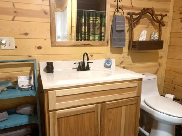 Log cabin bathroom sink with cream countertop, a mirror above it, blue hanging towels and a white toilet.