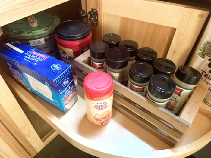 Wooden lazy Susan showing 9 spice jars with black lids, coffee and filters and a blue hot chocolate mix box
