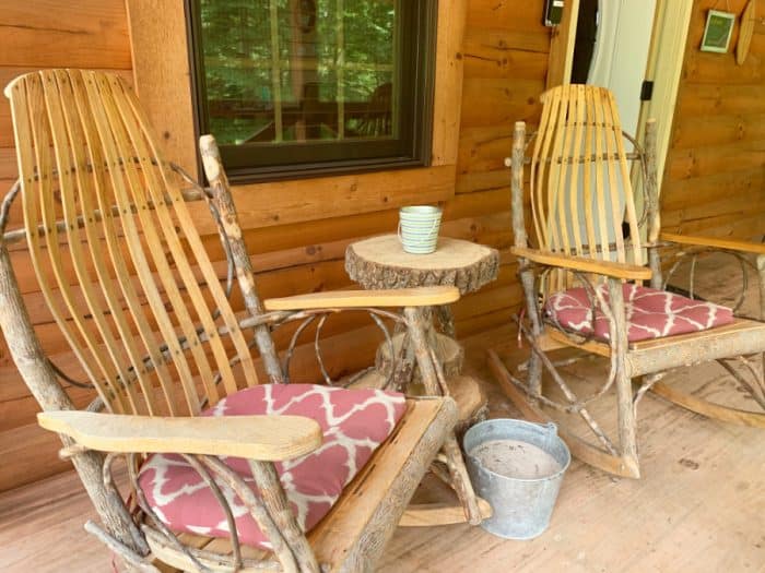 Two log and wooden rocking chairs on a porch with a log table and small candle jar on top and a pail of sand on the ground