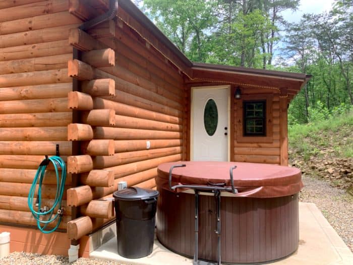 Log cabin with a green garden hose hanging on it, a black trash can, a brown covered hot tub and white door to the cabin.
