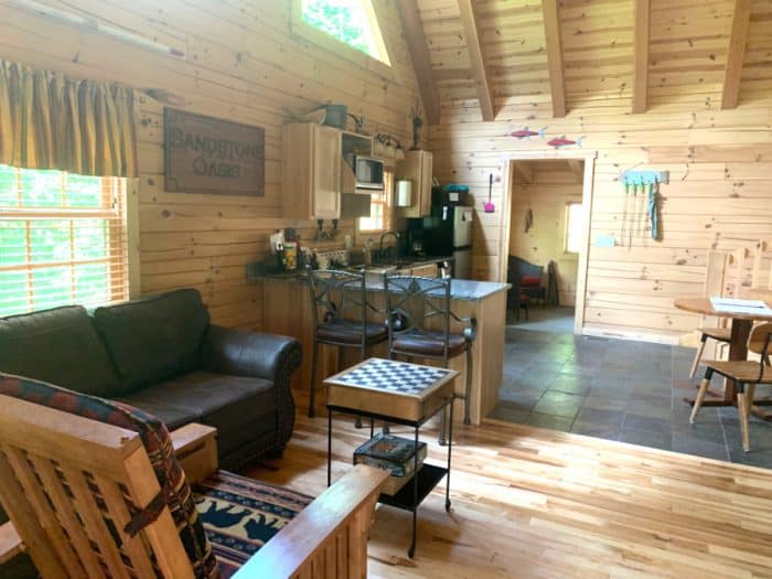 Cabin with light wooden walls, a chair, brown leather couch, kitchen and island with two bar stools and a table to the right